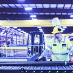 Marmon Keystone A worker in a high-visibility jacket and hard hat operates machinery on a conveyor belt in a large, well-lit industrial warehouse. Shelves of tube, pipe, and bar metals highlight the busy supplychain distributor environment.