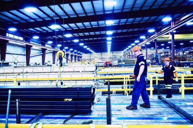 Marmon Keystone Two workers in safety gear walk near stacked metal pipes inside a brightly lit, large industrial warehouse with high ceilings, yellow safety railings, and an extensive metals supplychain.