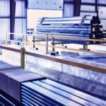 Marmon Keystone Stacks of metal pipes, tubes, and rectangular bars are organized inside a large industrial warehouse with concrete floors and yellow railings. Several cardboard boxes sit on top of some metal bars, ready for supply chain distribution.