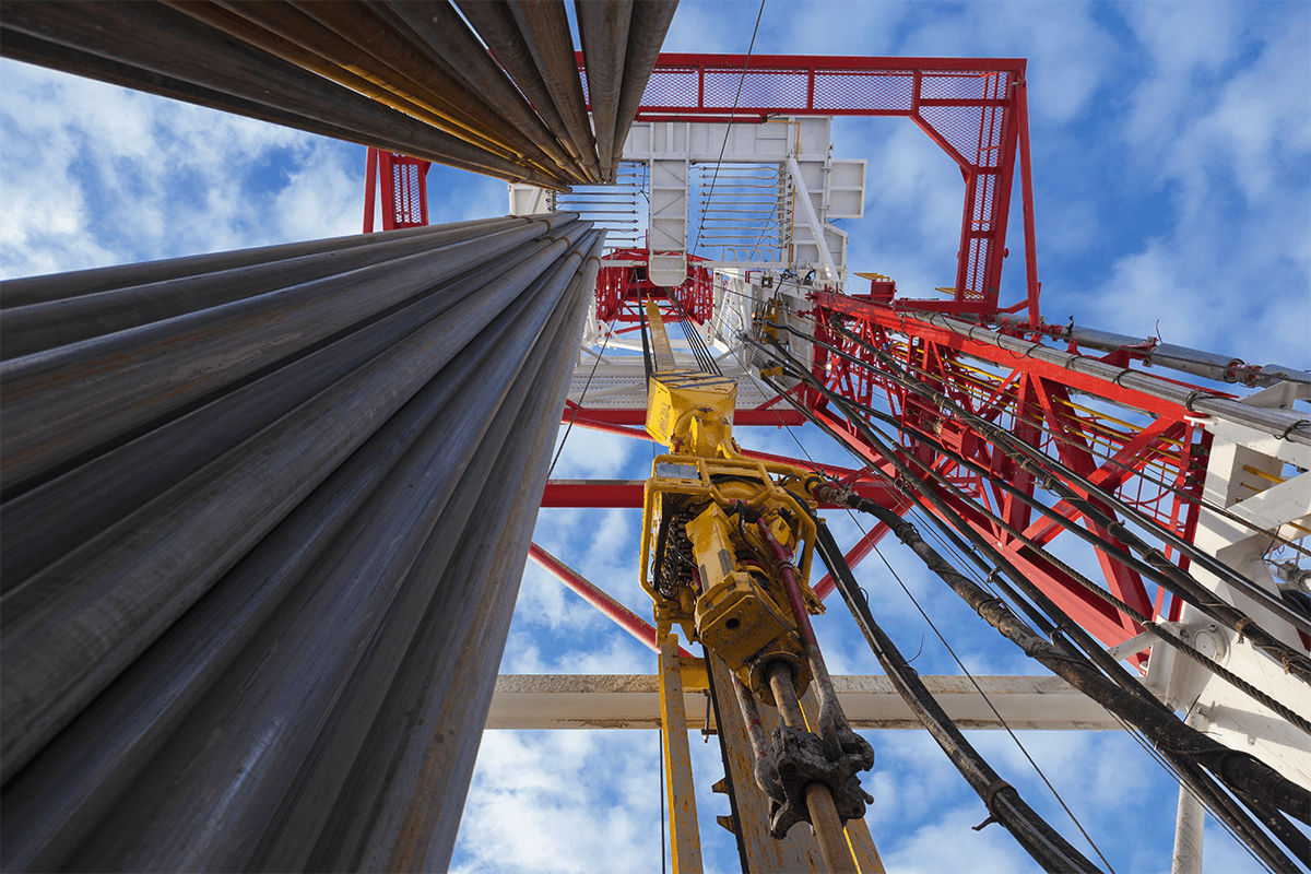 Marmon Keystone Low-angle view looking up at an oil drilling rig against a blue sky with clouds, featuring metal pipes supplied by a leading distributor, machinery, and red-and-white structural components essential to the supply chain.