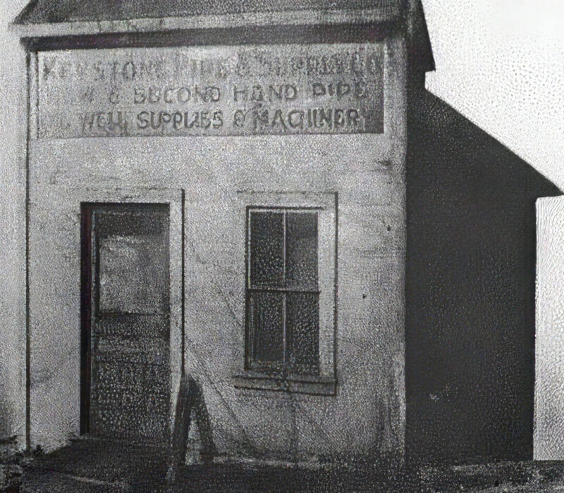 Marmon Keystone A black-and-white photo of an old building with a faded sign reading, “SECOND HAND BLDG. / IRON SUPPLIES & MACHINERY.” Once a metals and tube distributor, its door and window stand as reminders of its place in the local supply chain. The walls show signs of age.