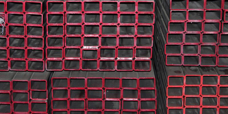 Marmon Keystone Stacks of rectangular hollow structural steel sections with red-painted edges are arranged in rows. The text above reads "Marmon/Keystone" and below reads "IN STOCK HSS metals distributor".