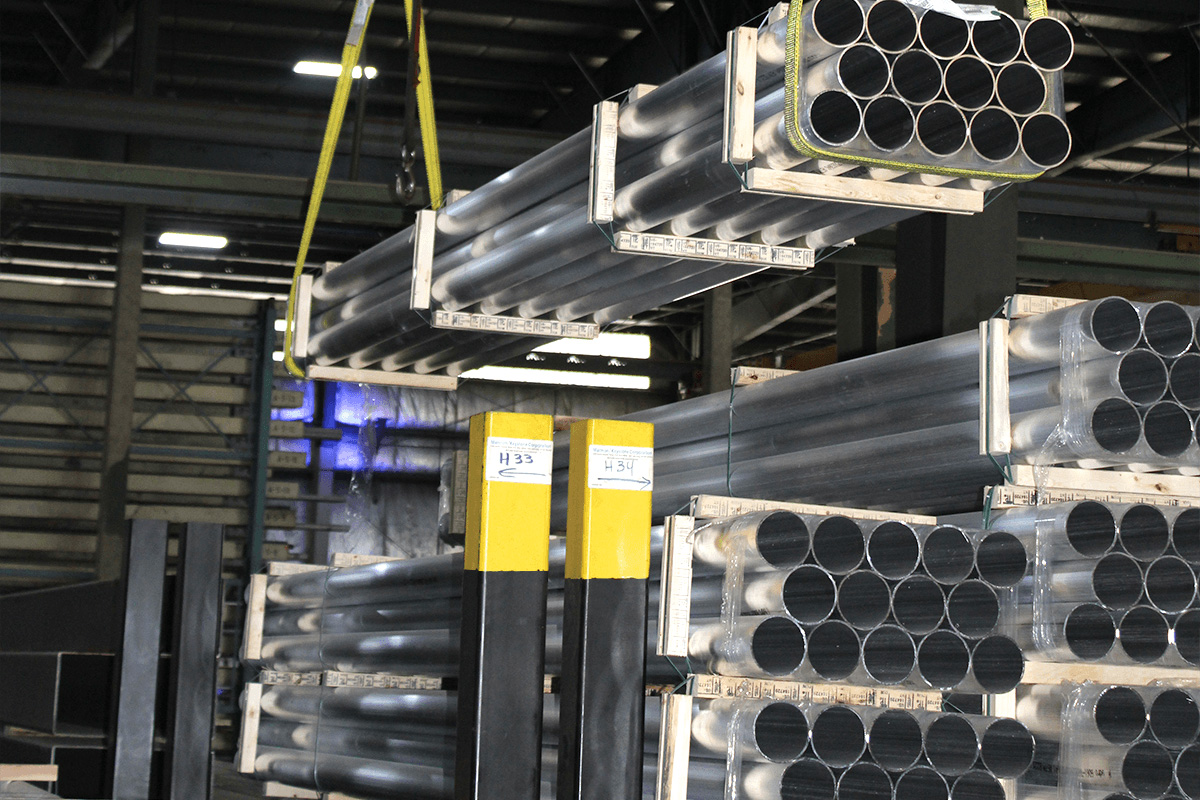 Marmon Keystone Bundles of black metal tube pipes are stacked on pallets in an industrial warehouse. A crane lifts one bundle while others are stored on the ground and metal shelves. Yellow safety posts are visible in the foreground.